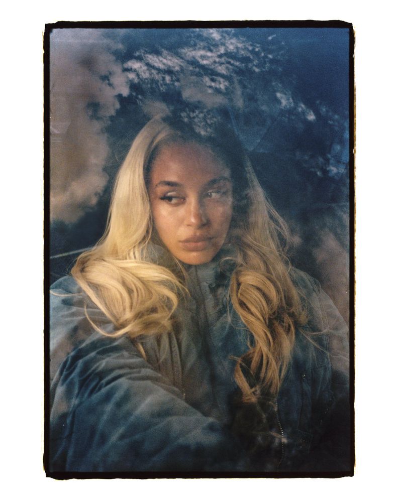 Portrait of a woman with long blonde hair, wearing a blue jacket, with a cloudy sky background, in a portrait orientation.