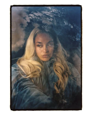 Portrait of a woman with long blonde hair, wearing a blue jacket, with a cloudy sky background, in a portrait orientation.