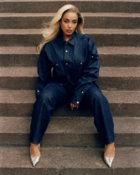 Singer Jorja Smith sitting on concrete stairs wearing a dark denim jacket, matching jeans, and silver pointed-toe heels.