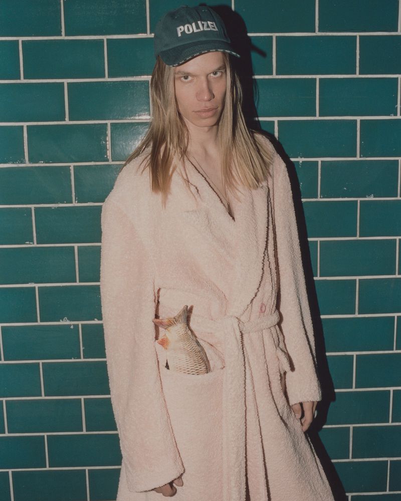 Person wearing a pink robe and a cap in front of a green tiled wall. The robe's pocket holds a fish.
