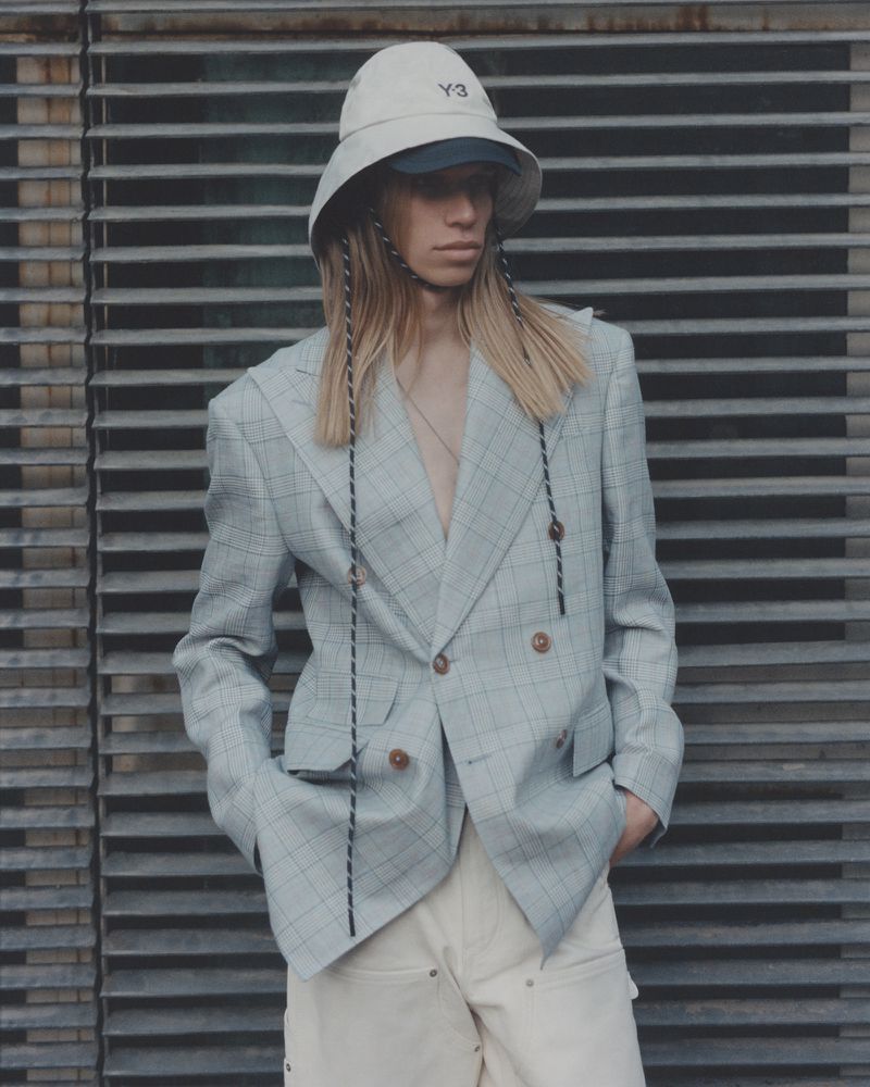Fashion-forward portrait of a young adult male model wearing a bucket hat, a plaid blazer, and beige pants, posing in front of metal grates.