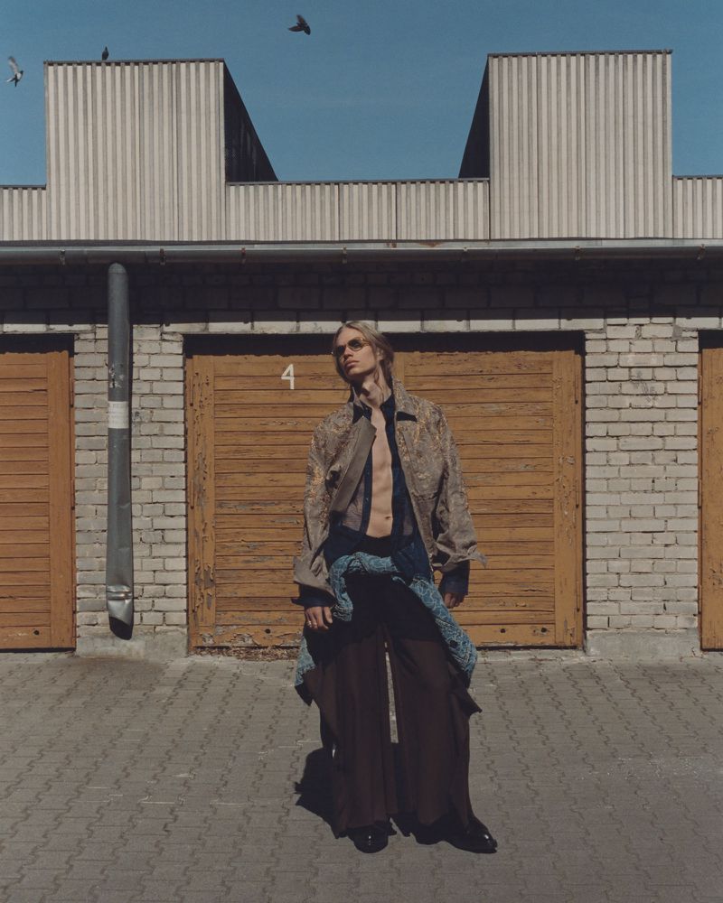 A slim blonde male fashion model stands outside wearing a patterned jacket, dark baggy pants, and sunglasses against garage doors.