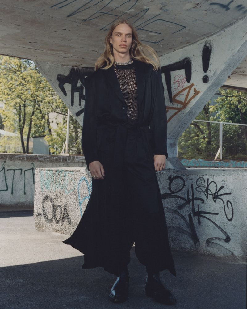 Model with long blonde hair wearing a black jacket and long skirt standing under a concrete bridge with graffiti