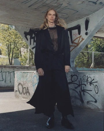 Model with long blonde hair wearing a black jacket and long skirt standing under a concrete bridge with graffiti