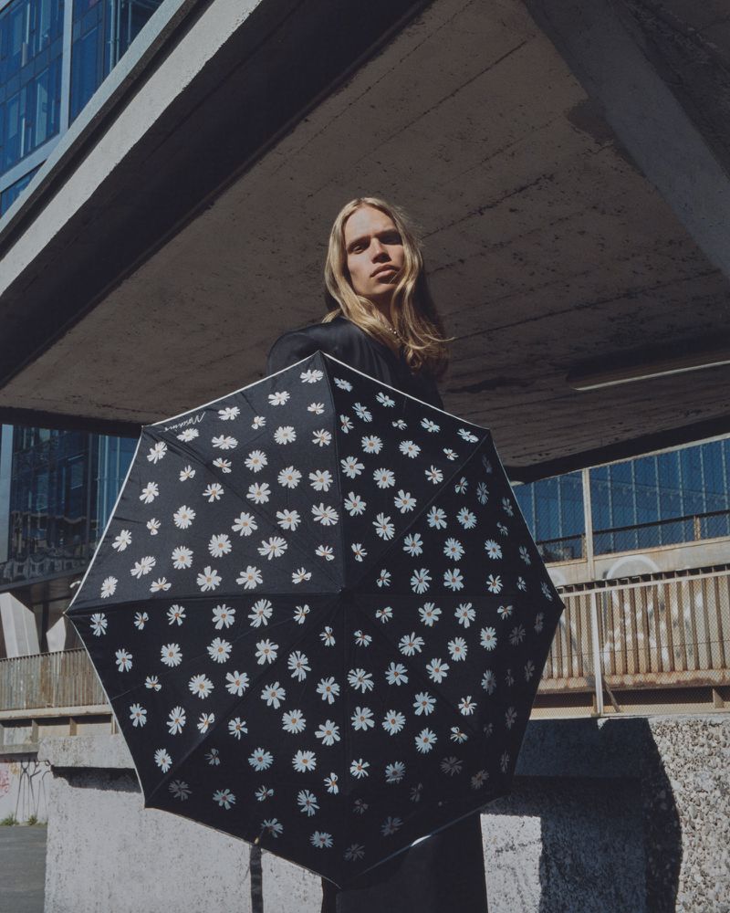 Young adult poses under concrete structure with a floral-patterned umbrella in an urban setting with light, neutral color palette.