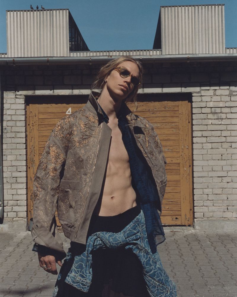 Young adult male poses in front of a brick building wearing a jacket, black shorts, and sunglasses in an outdoor setting.