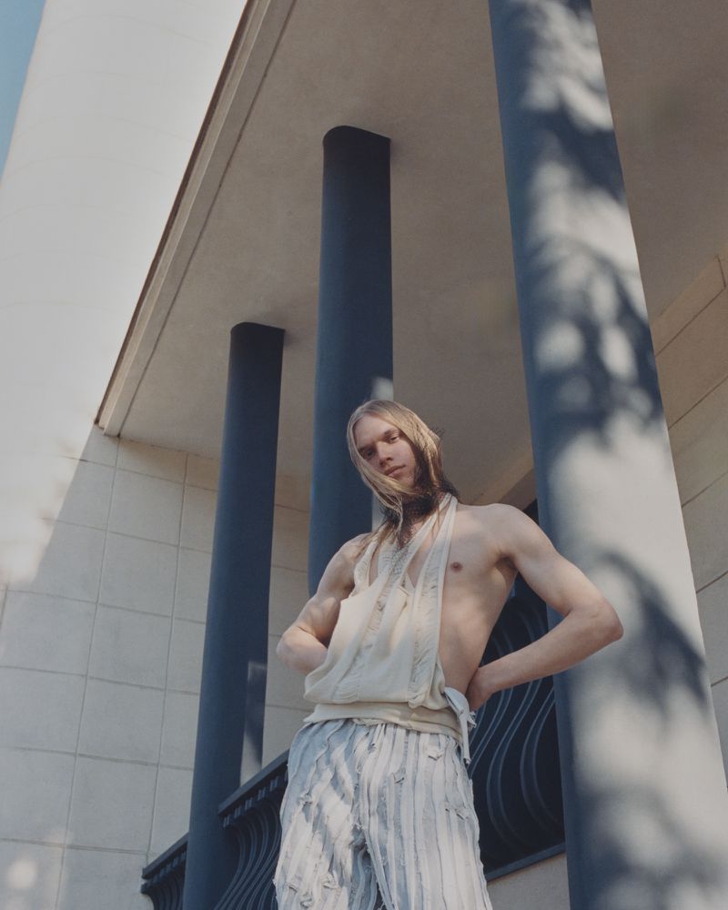 Blond male model poses outdoors in deconstructed casual wear against a modern building with columns on a sunny day in an eye-level shot.