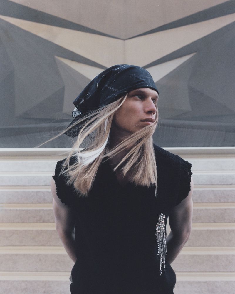 Young adult with a black bandana and blonde hair poses in front of grey geometric background and steps, wearing a black sleeveless top.