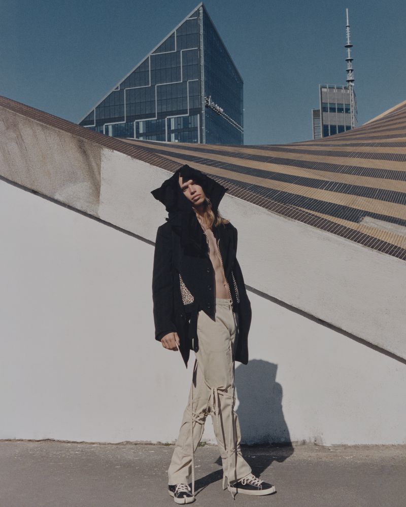 Model Bartek Niewiadowski wears a black hooded jacket and beige strapped trousers against geometric modern architecture.