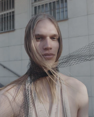 Close-up portrait of a fair-skinned adult with blond hair, accessorized with a black fishnet scarf, set against a neutral background.