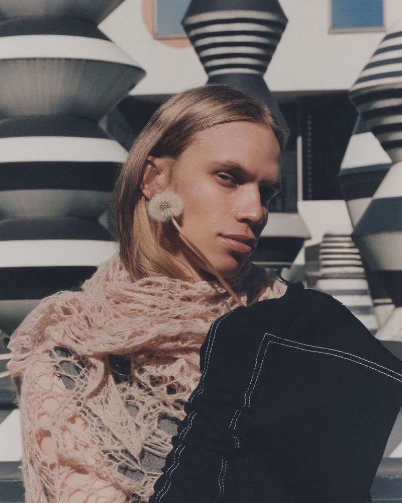 Portrait of androgynous model with blonde hair wearing a pale pink knit shawl and holding a dandelion against geometric patterned background.
