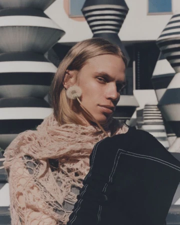 Portrait of androgynous model with blonde hair wearing a pale pink knit shawl and holding a dandelion against geometric patterned background.