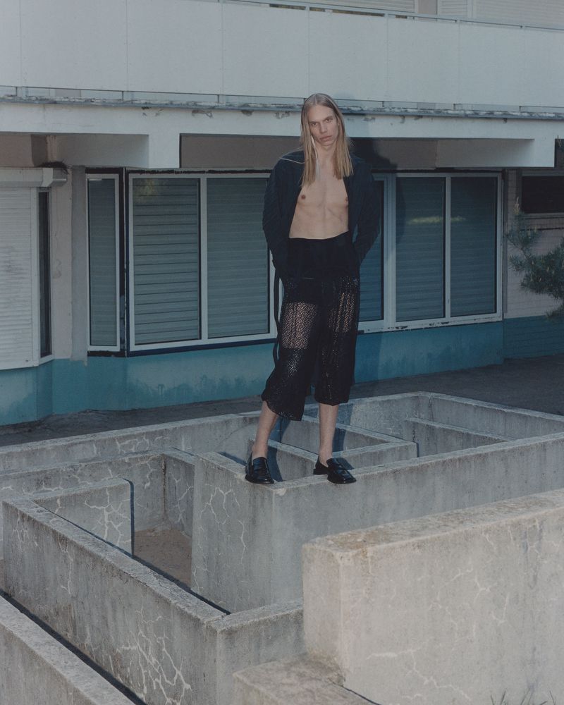 A tall, fair-haired adult stands in a concrete maze, wearing a black jacket and lace capri pants, posing in a fashion editorial.