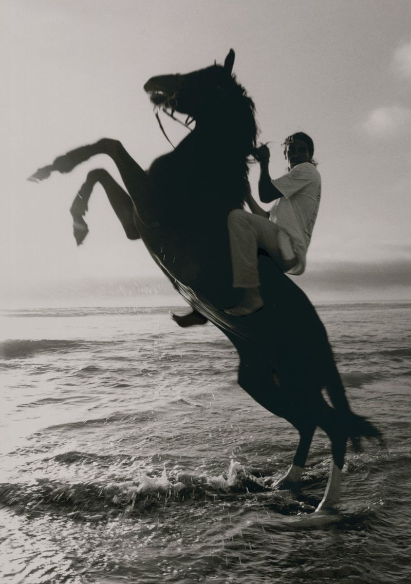 A man rides a rearing dark horse along a shoreline as waves crash in this monochrome outdoor scene captured in a medium shot.