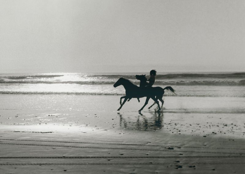 Black and white shot of person riding a horse on the beach with ocean waves in the background. Equestrian scene on the sand.