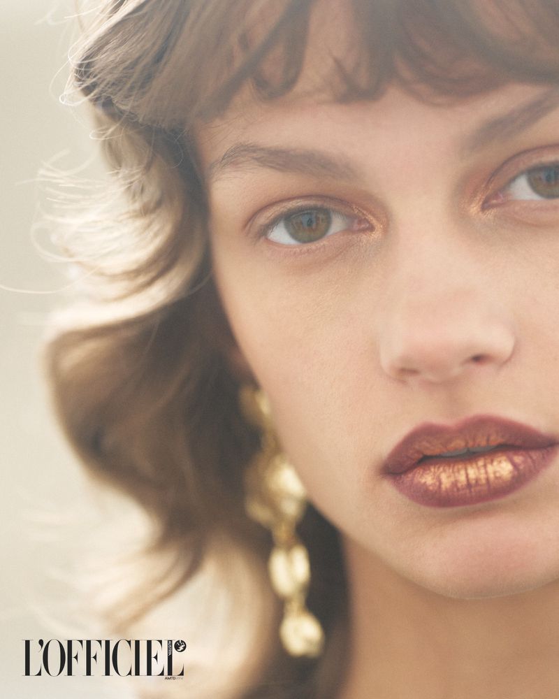 Close-up portrait of a young woman with fair skin, brown eyes, and gold lipstick, wearing gold earrings, posing for a beauty shot.
