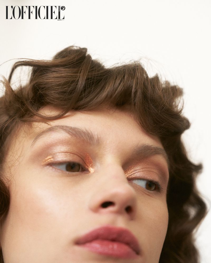 Close-up portrait of a brunette woman with golden eyeshadow and glossy lips against a clean white background, looking to the side.