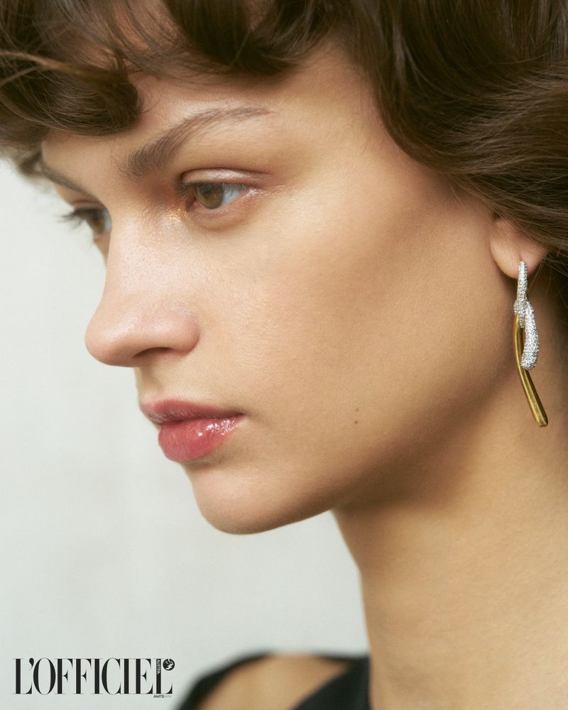 Close-up shot of an adult female model with short brown hair wearing a gold and silver earring in a studio with soft, diffused lighting.