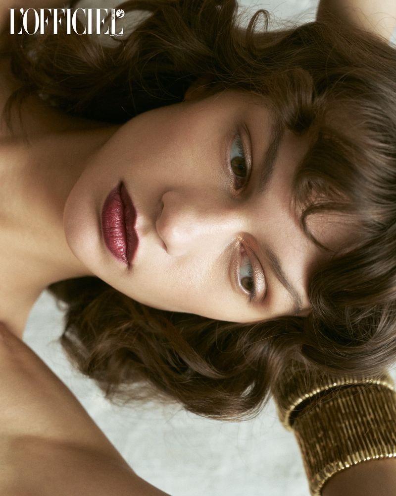Close-up portrait of a young woman with curly brunette hair and dark red lipstick, wearing a gold cuff bracelet. Beauty shot for L'Officiel.