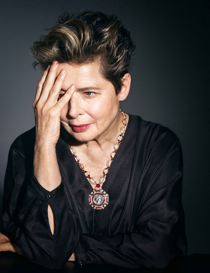 Mature woman with short blonde hair, wearing a black dress and necklace, poses in a close-up studio shot with a dark background.
