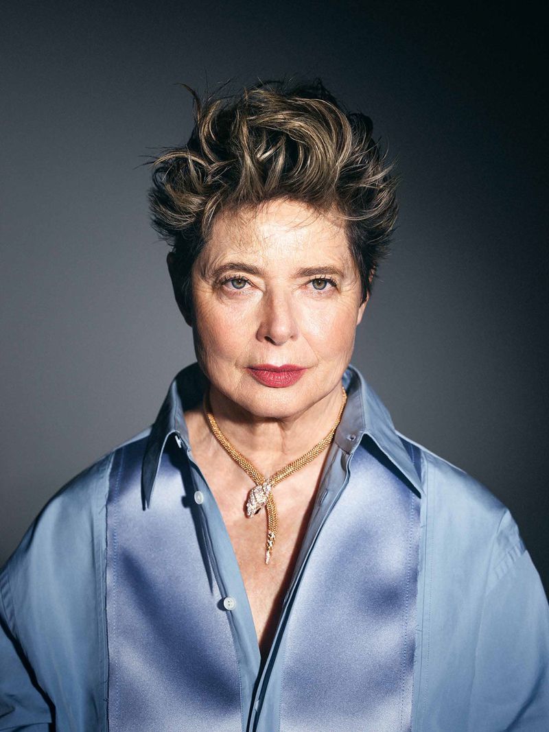 Portrait of Isabella Rossellini wearing an open light blue satin shirt and a gold snake necklace against a grey studio backdrop.