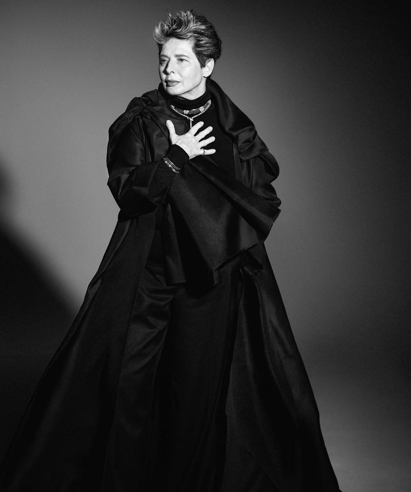 Black and white portrait of a woman wearing a long black dress and a large, ornate necklace in a photo studio setting.