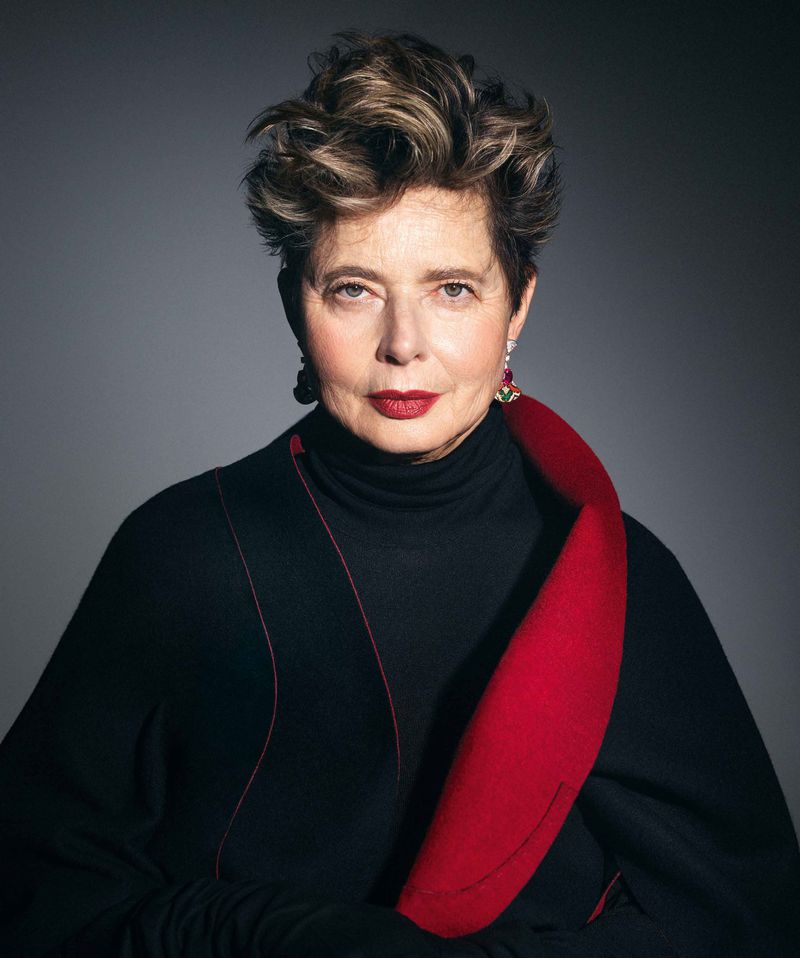 Portrait of an adult female with short, wavy hair, wearing a black top with a red collar, set against a grey backdrop.