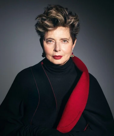 Portrait of an adult female with short, wavy hair, wearing a black top with a red collar, set against a grey backdrop.
