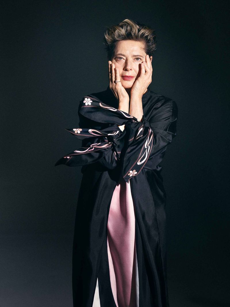 Adult woman with short hair poses in a black robe with hands on her cheeks against a dark gray background in a studio portrait.
