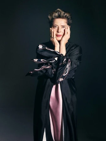 Adult woman with short hair poses in a black robe with hands on her cheeks against a dark gray background in a studio portrait.