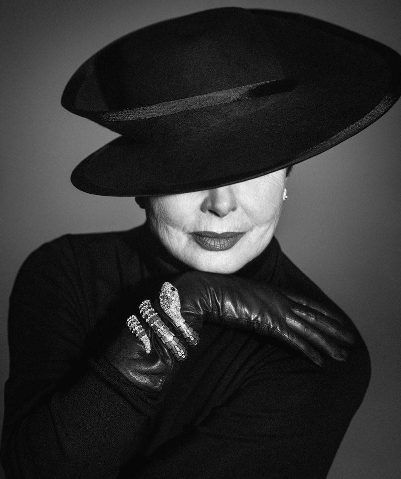 Monochrome portrait of a mature woman with a wide-brimmed hat, black turtleneck, leather gloves, and a striking snake ring against a gray background.