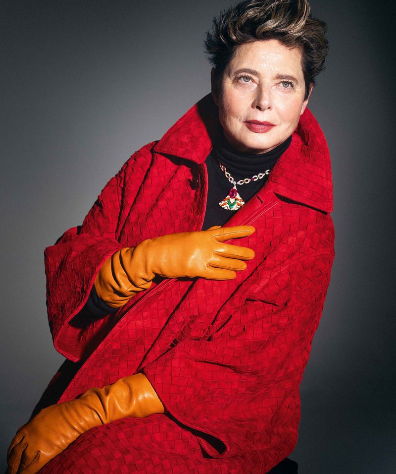 Older woman in a red patterned coat and tan leather gloves poses in a studio, looking at the camera with a neutral facial expression.