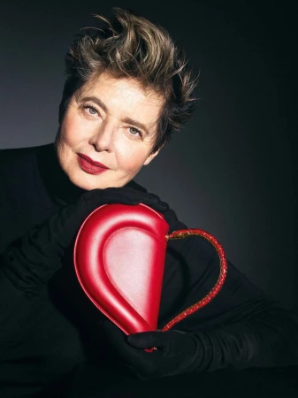 Isabella Rossellini wearing black gloves and holding a red heart-shaped leather clutch against a dark studio background.