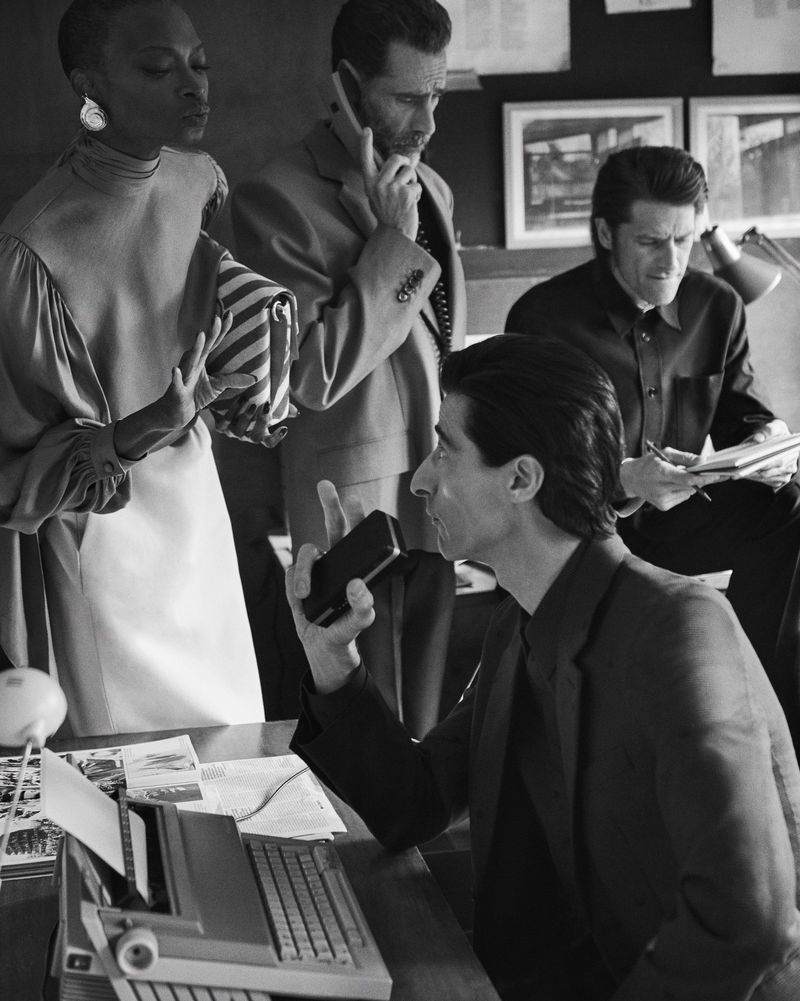Group of four adults in an office setting, one woman standing, three men, one on the phone, one holding a notebook, and a man at a typewriter.