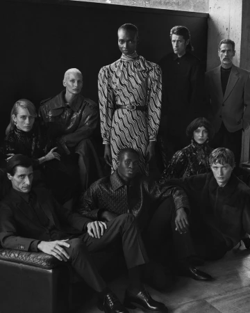 A group of nine fashion models poses indoors with black and white palette and artful composition, showcasing clothing and unique hairstyles.