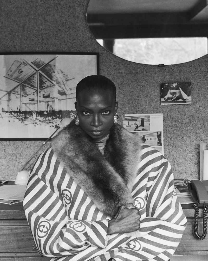 Black and white portrait of a Black woman with a shaved head wearing a striped jacket with fur collar and large earrings indoors.