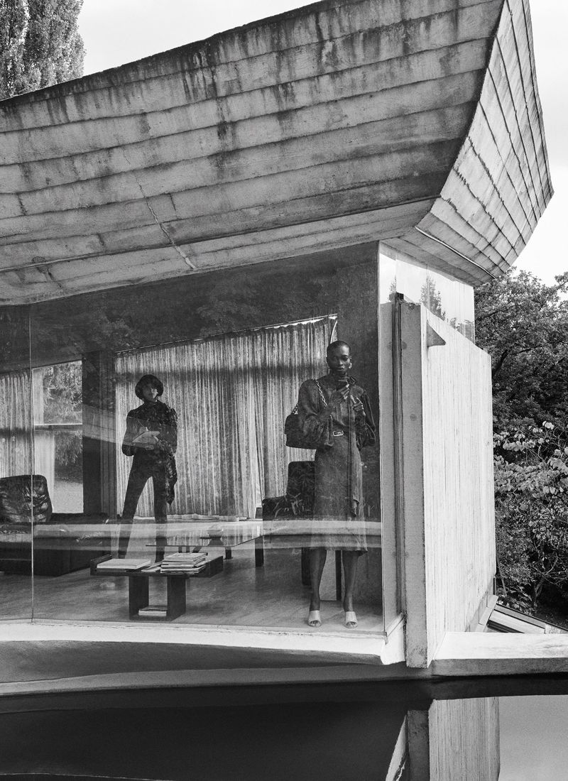 Black and white fashion shot of two women standing in a glass modernist home, reflected in a pool of water in a natural setting.