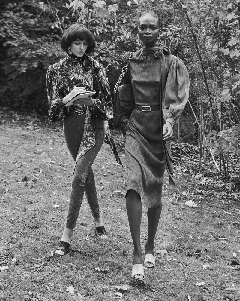 Two women in vintage-style dresses walk on a grassy path lined with foliage, one writing in a notebook, showcasing fashion in nature.