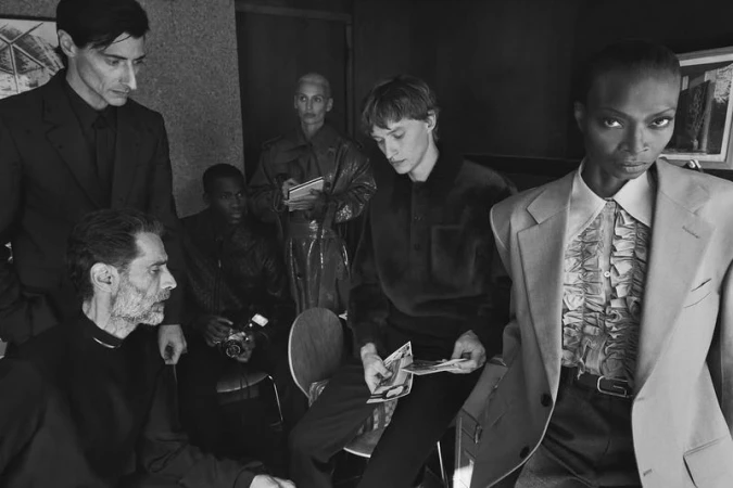 Group of seven fashionably dressed people in an indoor setting. A woman in a blazer looks at the camera, while others are seated or standing.