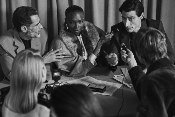 Black and white image of a Black woman sitting at a table being interviewed by two people with microphones and photographers around.