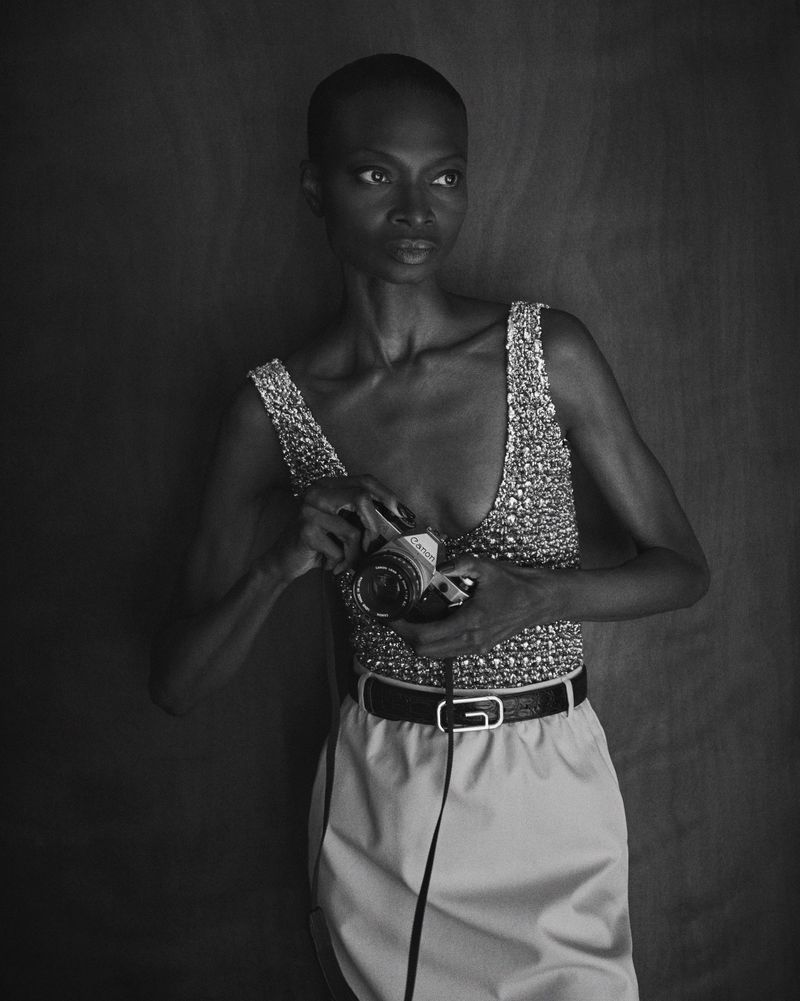 Black and white portrait of an adult female holding a vintage camera, wearing a shiny top and skirt, posing in a photo studio.