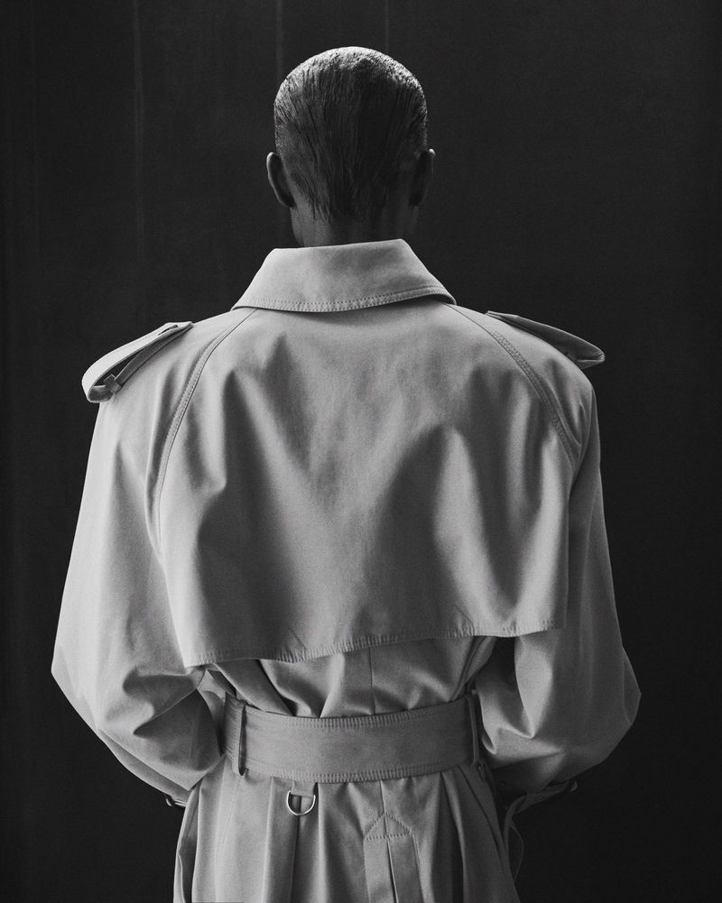 Back view of a person wearing a tan trench coat against a dark backdrop. The coat features a classic design with epaulets and a belt.
