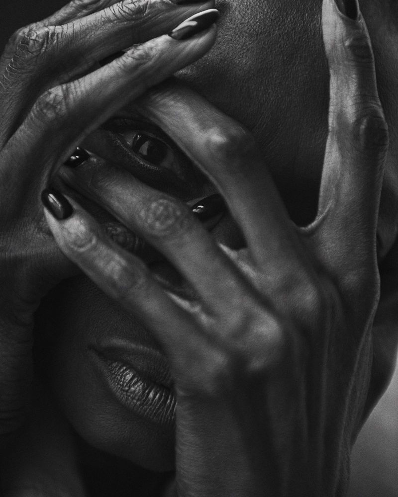 Monochrome close-up of a Black woman's face partially obscured by her hands with polished nails, emphasizing texture and skin tones.