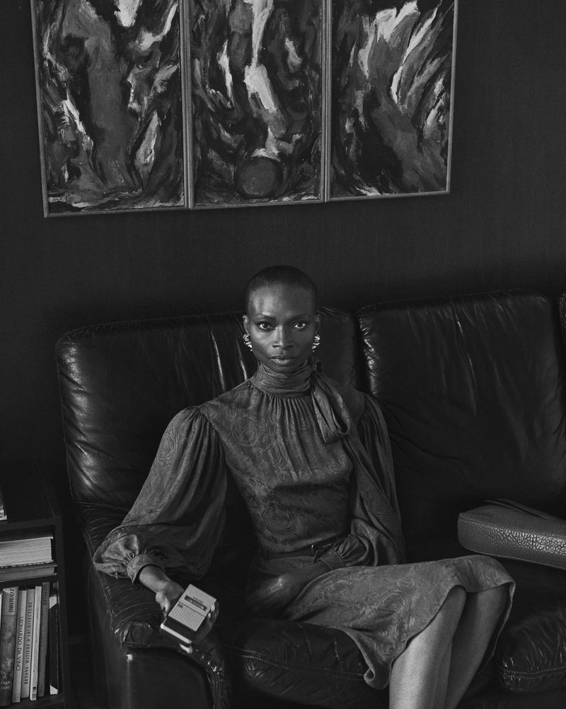 Black and white portrait of a Black woman sitting on a leather couch in front of artwork, wearing a patterned dress and large earrings.