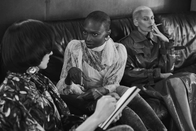 Black and white editorial image of three women seated on a leather sofa, with one person sketching in a notebook.