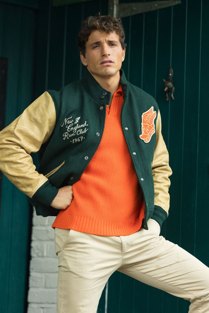 Portrait of a young adult male in a letterman jacket and orange knit shirt, posing outdoors with a neutral expression.