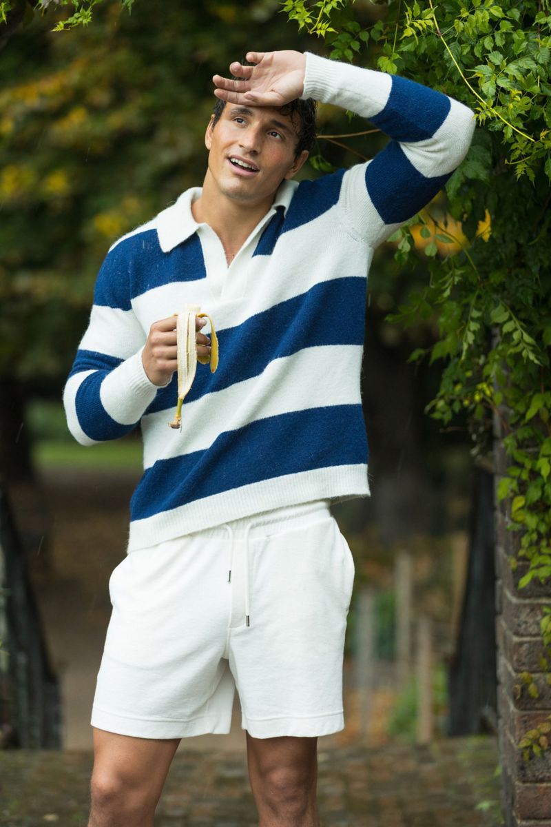 Young man in a striped polo shirt and white shorts stands outdoors eating a banana, with greenery in the background and dappled light.