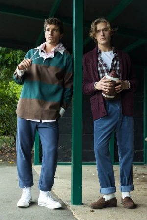 Two young men pose outdoors in vintage-inspired casual outfits, one holding a football, with a green foliage background.