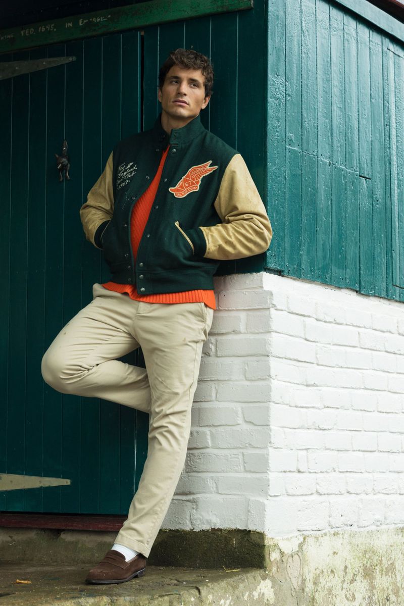 A stylish man in a varsity jacket leans against a green door and white brick wall, creating a fashion-forward, casual outdoor portrait.
