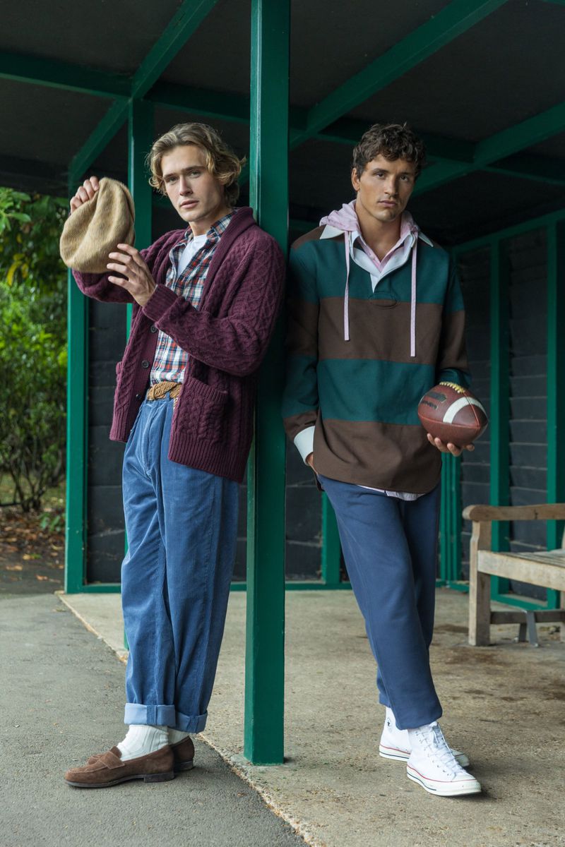 Two young men stand outdoors in a covered area, one holding a hat, the other an American football, wearing casual fall fashion.
